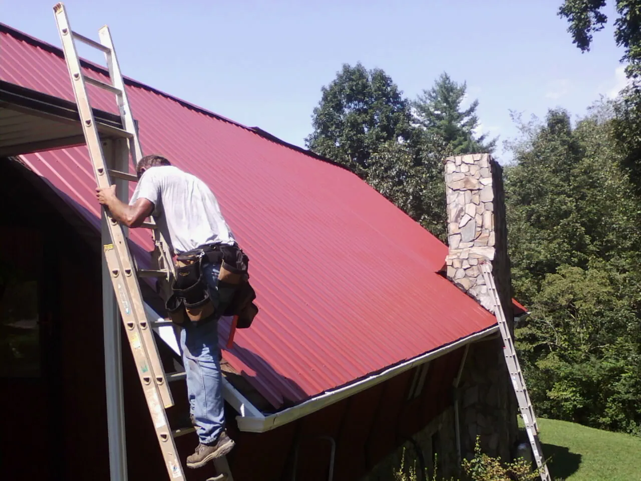 Roofer on a ladder beside a red metal roof and stone chimney — Soffit & Fascia Repair by Roofing Brothers in Lawson